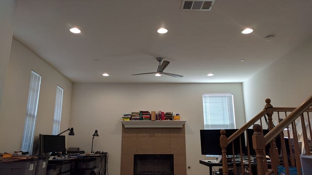 Modern living room with a ceiling fan, fireplace, books on the mantel, and two desks with computers.