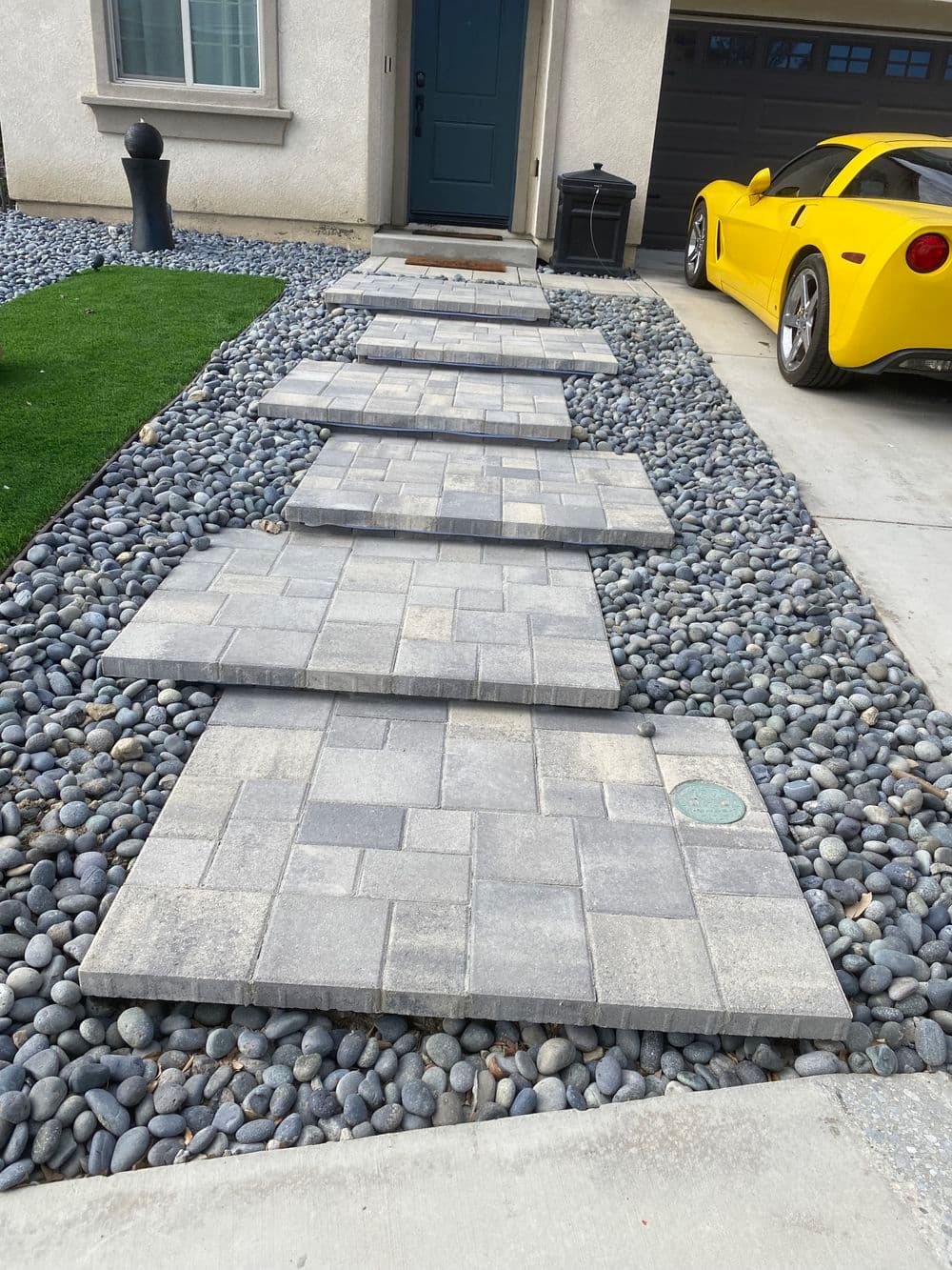 Stone walkway with gray pavers and yellow sports car beside a neatly landscaped yard.