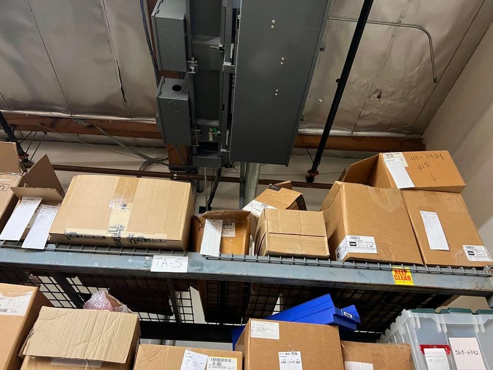 Storage area with stacked cardboard boxes on metal shelves, overhead equipment visible.