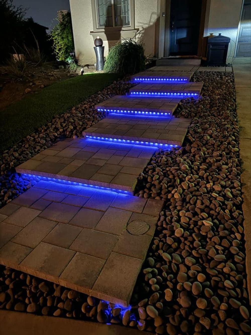 Illuminated blue stone pathway leading to a house entrance at night, surrounded by pebbles.