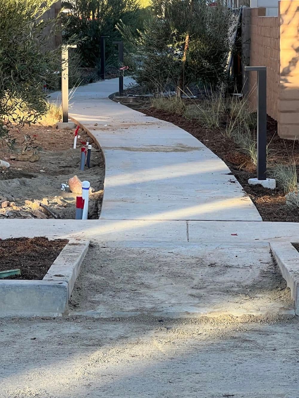 Curved concrete walkway surrounded by plants and construction elements in a landscaped area.